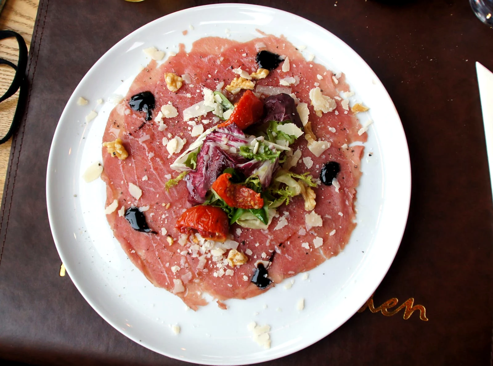 Carpaccio met Parmezaan, walnoten, salade en zongedroogde tomaat op een wit bord.