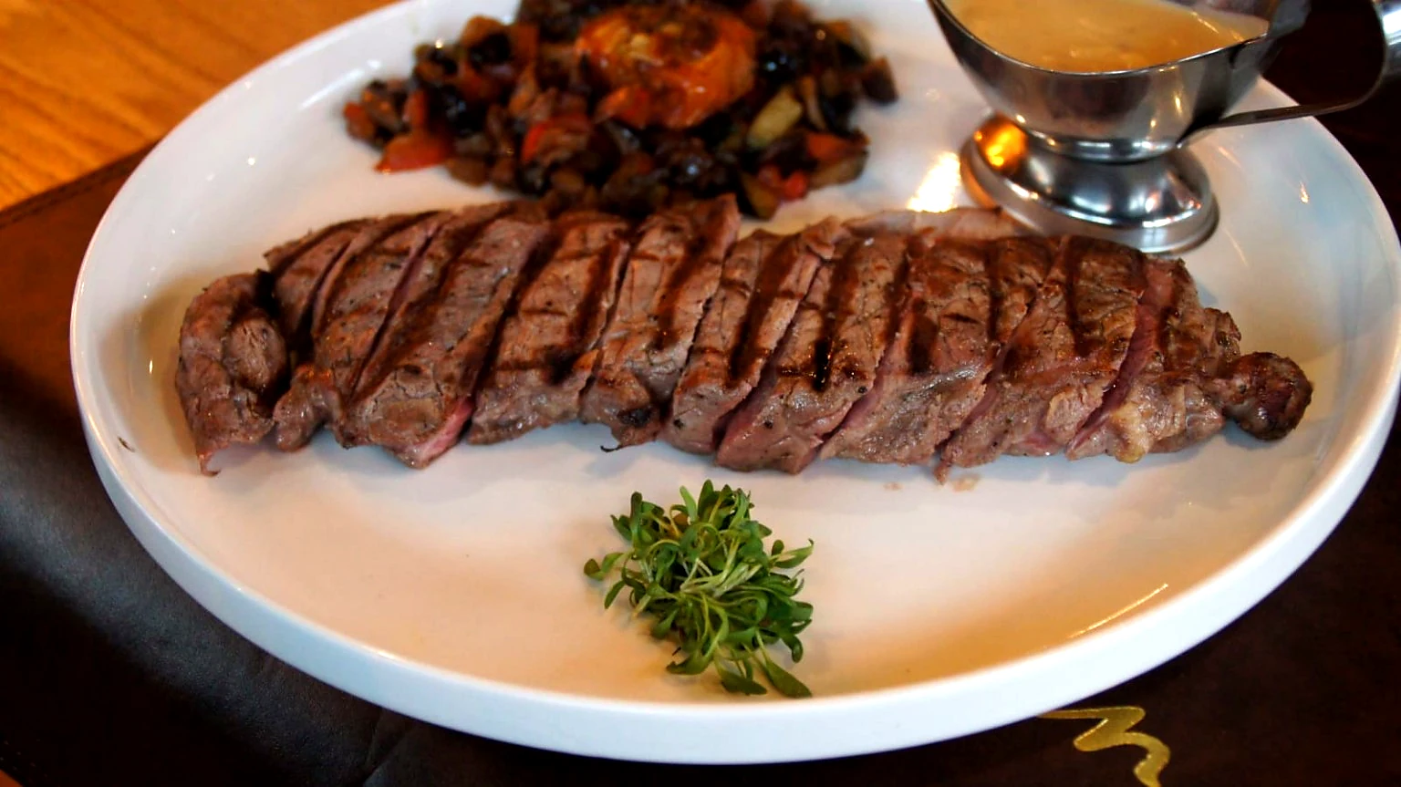 Gegrilde steak met groenten, saus en verse kruiden op een wit bord.