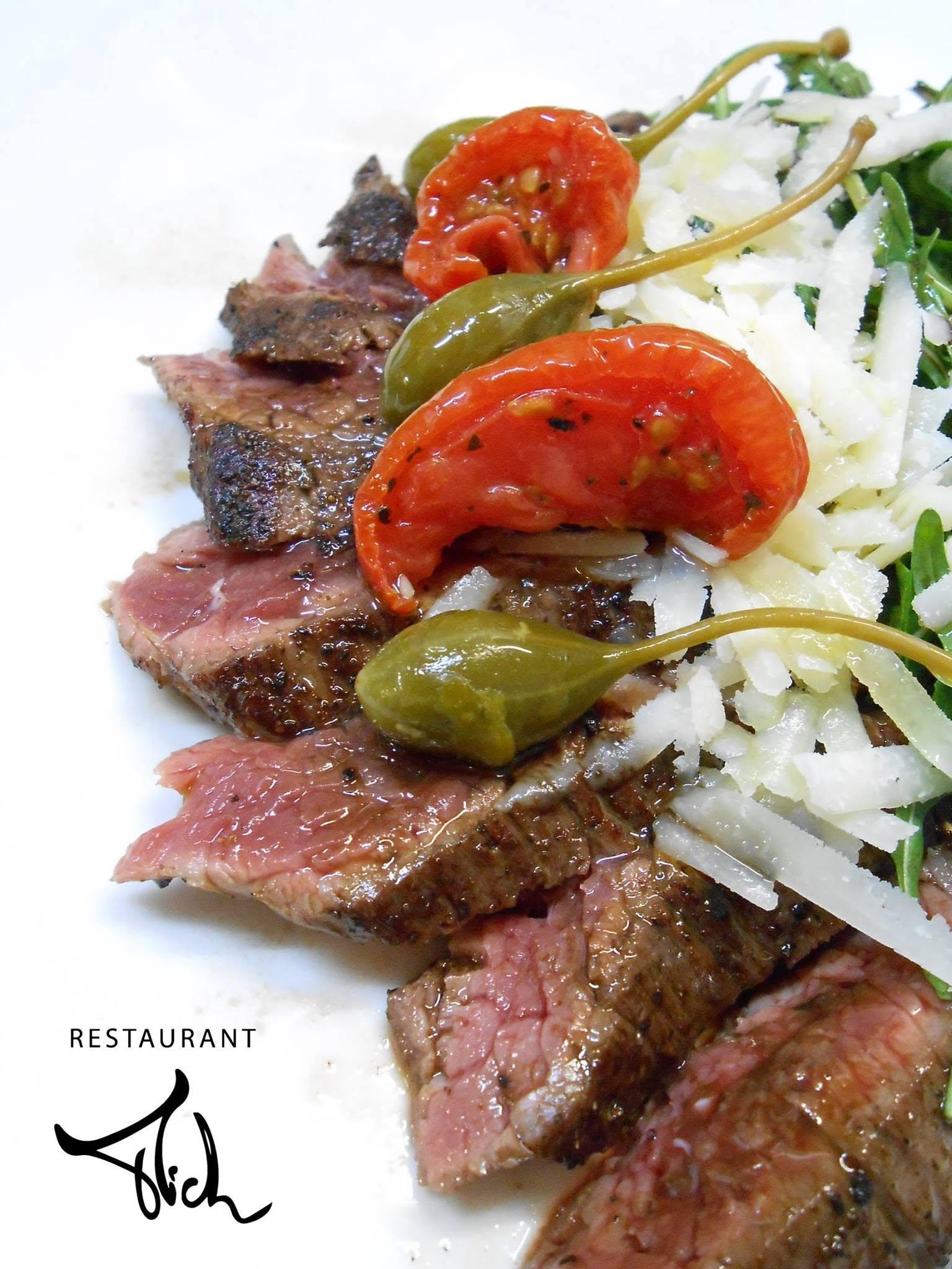 Steakgerecht met rucola, kaasschilfers, tomaat en kappertjes, afgewerkt met het Restaurant Mich-logo.