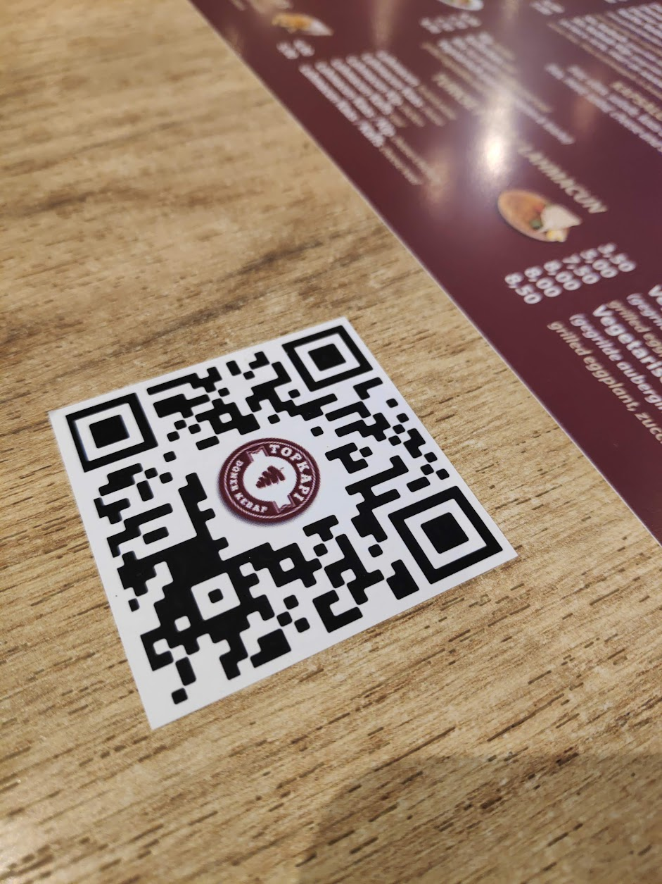 Authentieke Topkapi QR-sticker met het restaurantlogo op een houten tafel naast de menukaart.