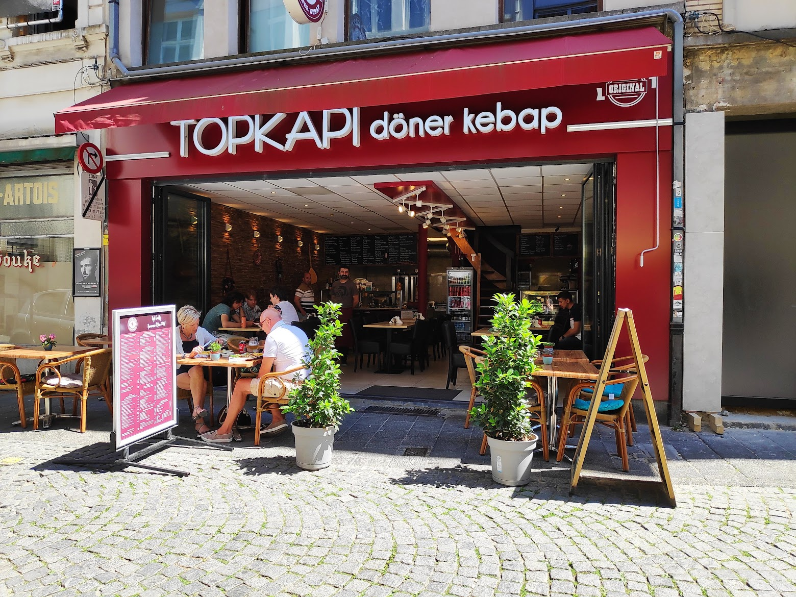Open gevel van Topkapi overdag met gasten op het terras, planten en medewerkers in de zaak.